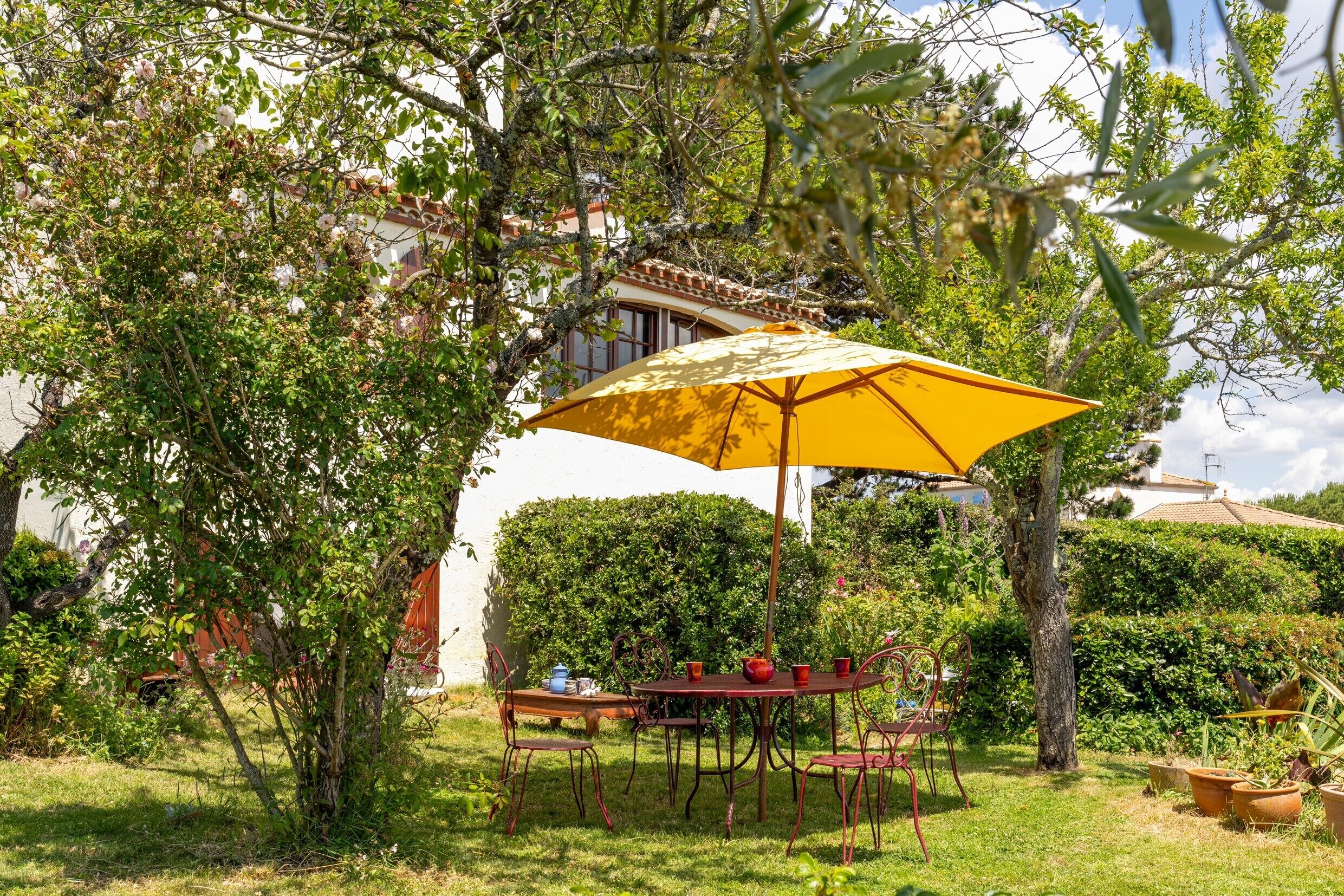 A patio with a table and chairs under a yellow umbrella, surrounded by greenery and a building in the background.