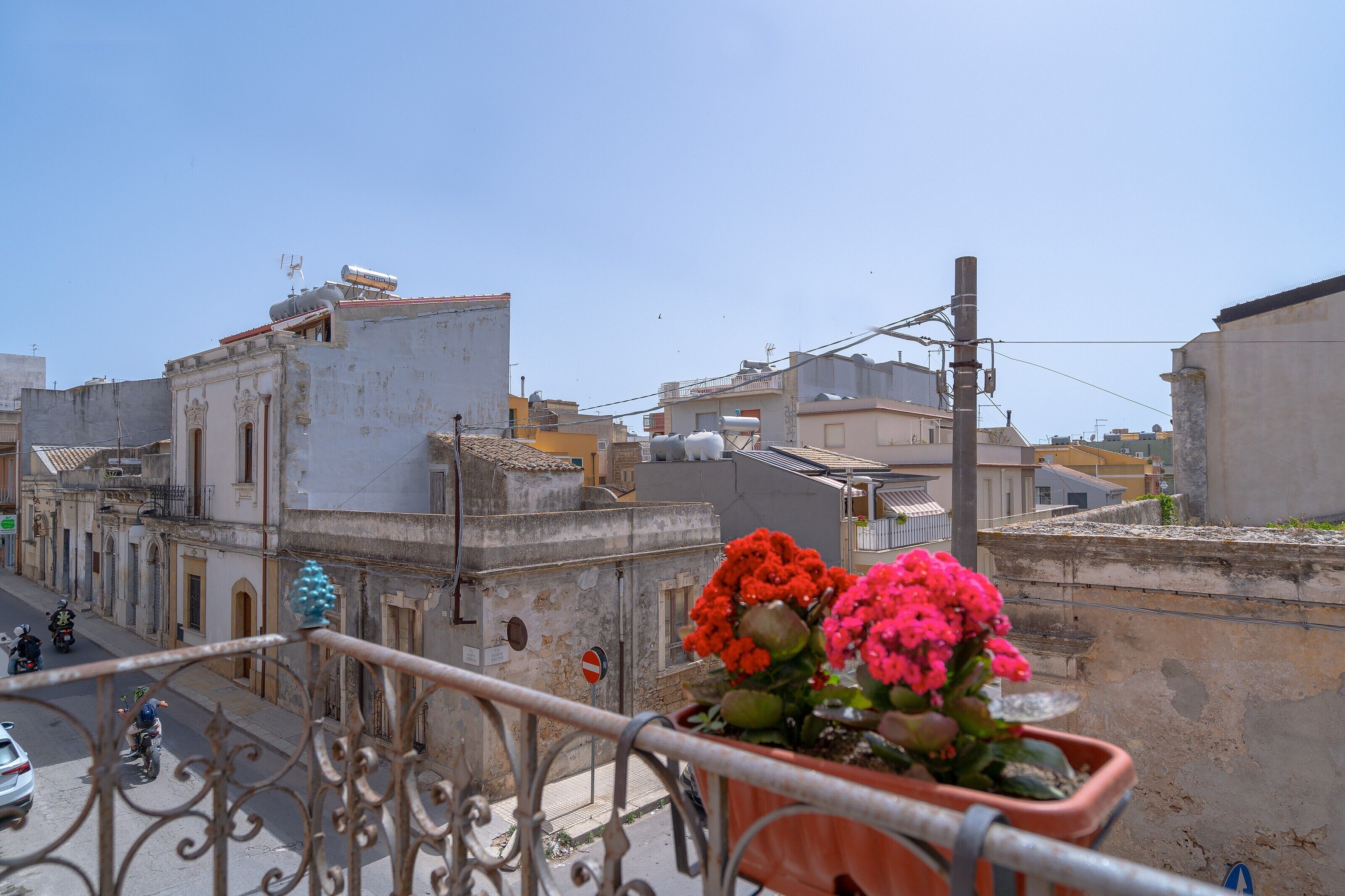 Un balcón con barandilla, una maceta con flores y vista a la calle con edificios y cielo despejado.