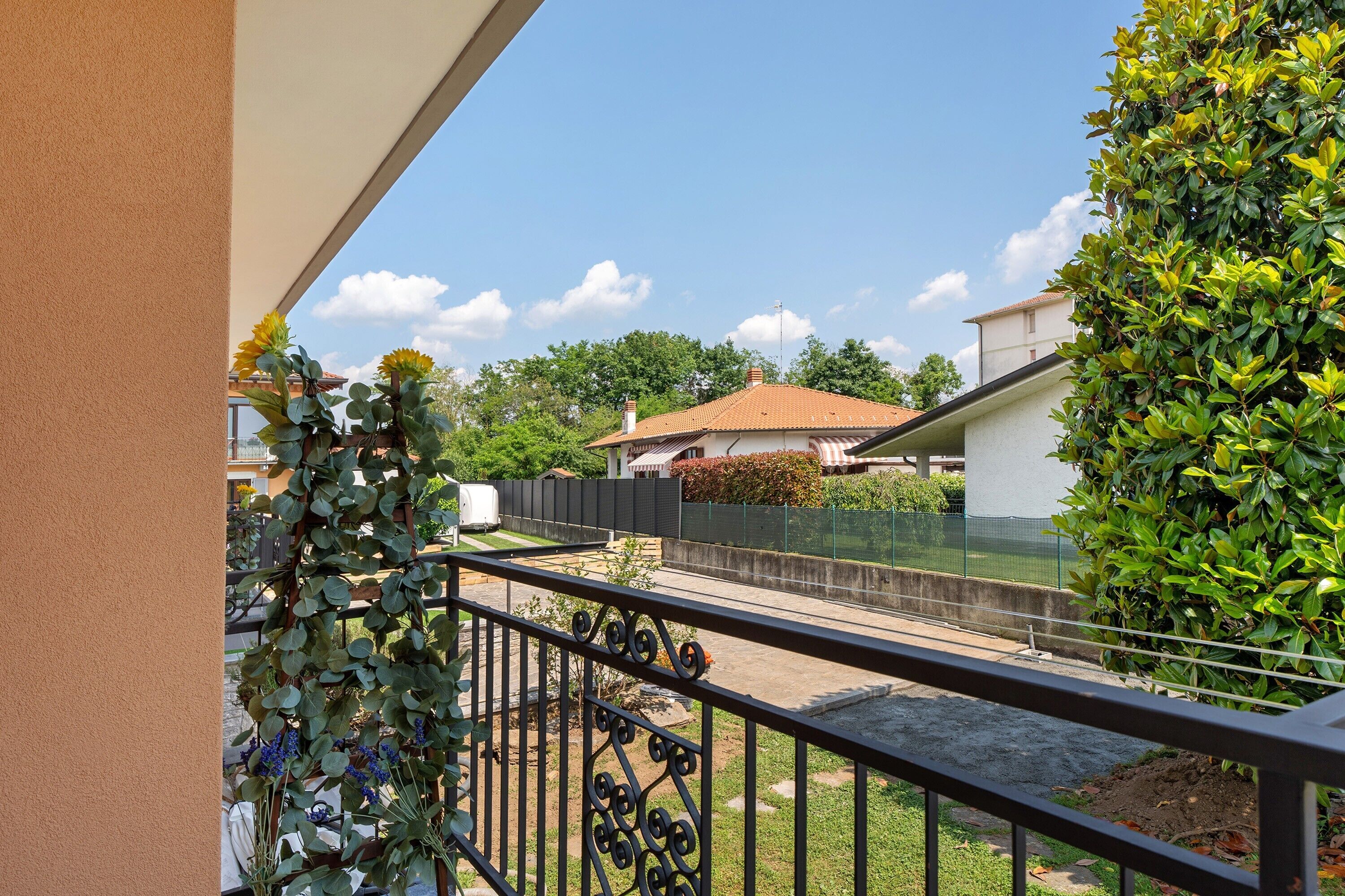 Ein Balkon mit Blick auf einen umzäunten Garten, ein Gebäude mit rotem Dach und viel Grün.