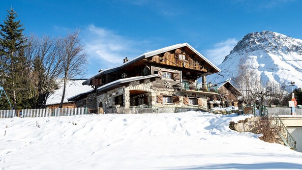 A wooden chalet with a stone foundation, a balcony, and a mountain backdrop. - Apartment 'Chalet -Les Aiguilles- Rdc' with Mountain View, Terrace and Garden (Albiez-Montrond)