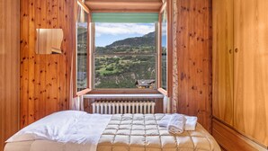 Ein Zimmer mit Holzwänden, einem Bett mit weißer Bettdecke und Kissen sowie einem Heizkörper unter einem Fenster mit Blick auf eine grüne Landschaft.