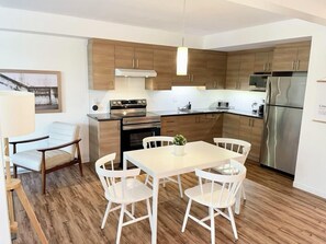 A modern kitchen with wooden cabinets, a stainless steel refrigerator, and a dining area with white chairs and a table.