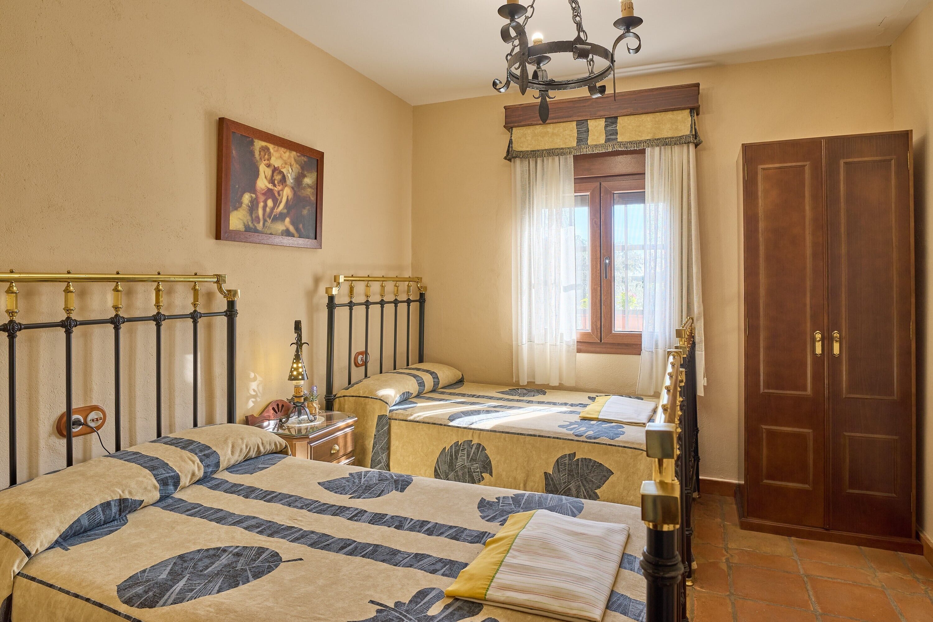 A hotel room with two beds, a chandelier, a painting on the wall, a window with curtains, and a wooden door.