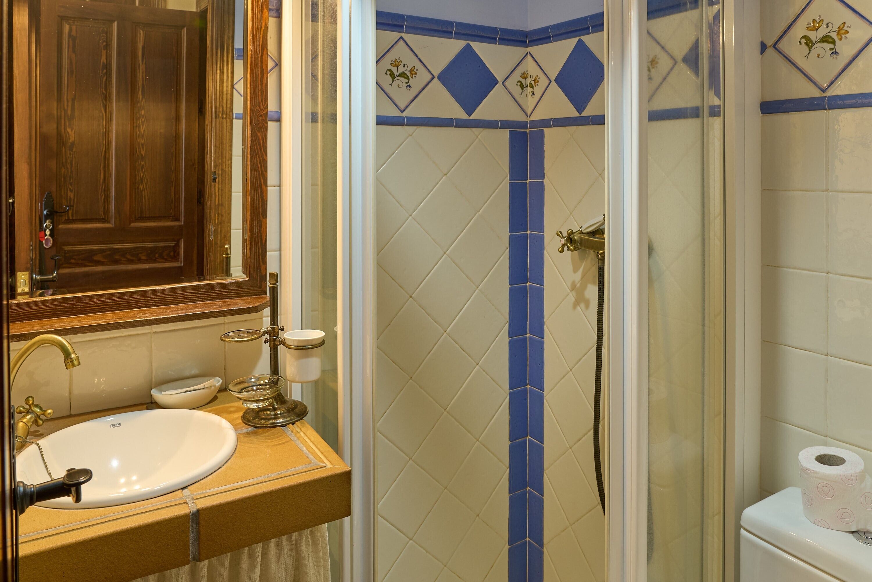 A bathroom with a wooden door, a sink, a mirror, a shower with blue tiles, and a toilet.