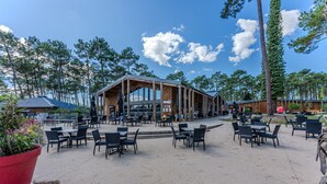 Un espace extérieur aménagé pour les repas, avec des tables et des chaises, un bâtiment doté de grandes fenêtres et un paysage boisé en arrière-plan.