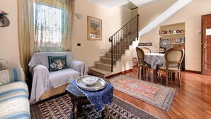 Una sala de estar con un sofá, una mesa de comedor, sillas, una escalera y una estantería.