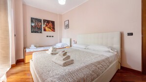 Habitación de hotel con una cama grande, una cama más pequeña, una mesita de noche y un calentador de pared, además de obras de arte en las paredes.