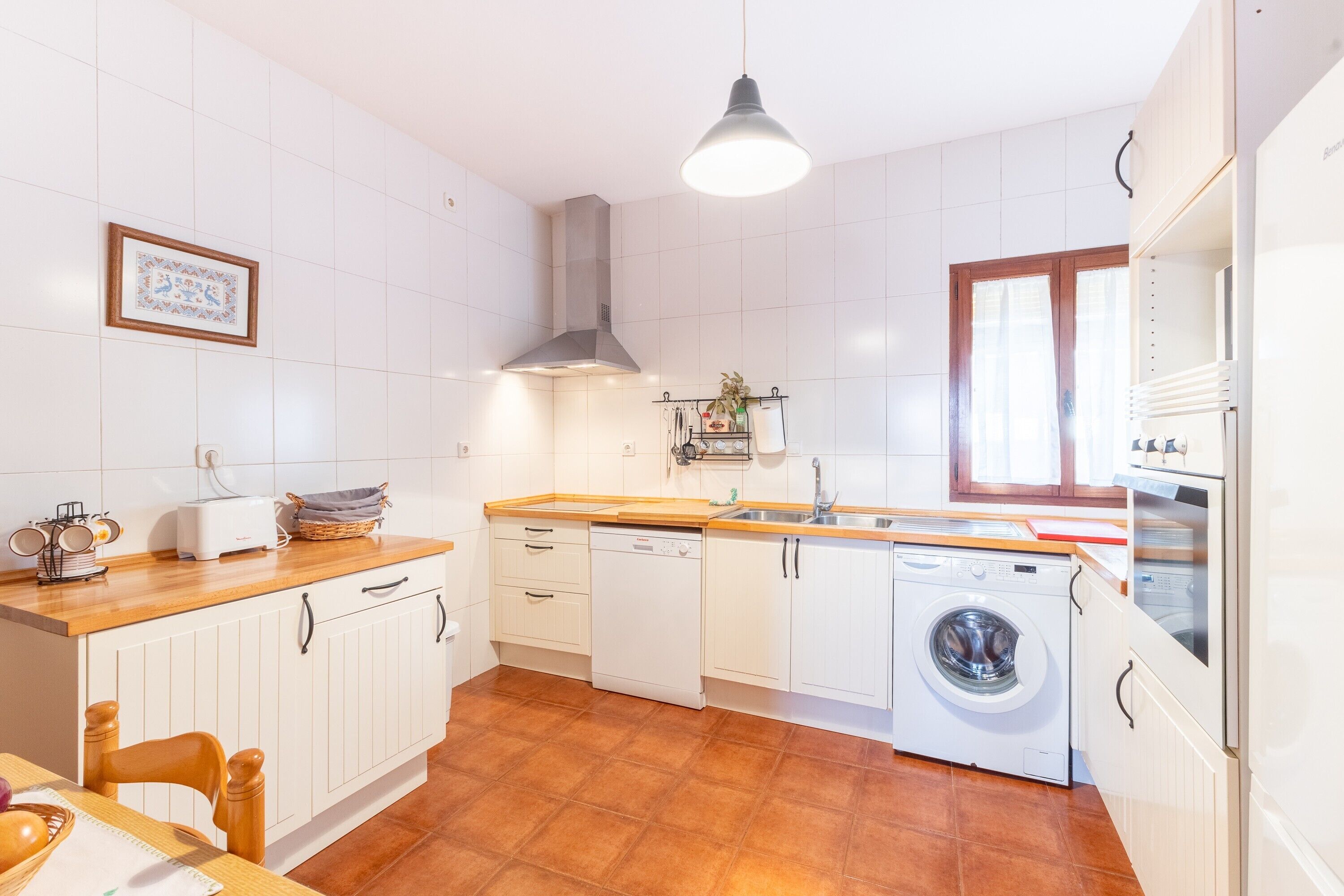 Une cuisine avec des meubles blancs, un lave-linge, un évier et une fenêtre avec des rideaux.