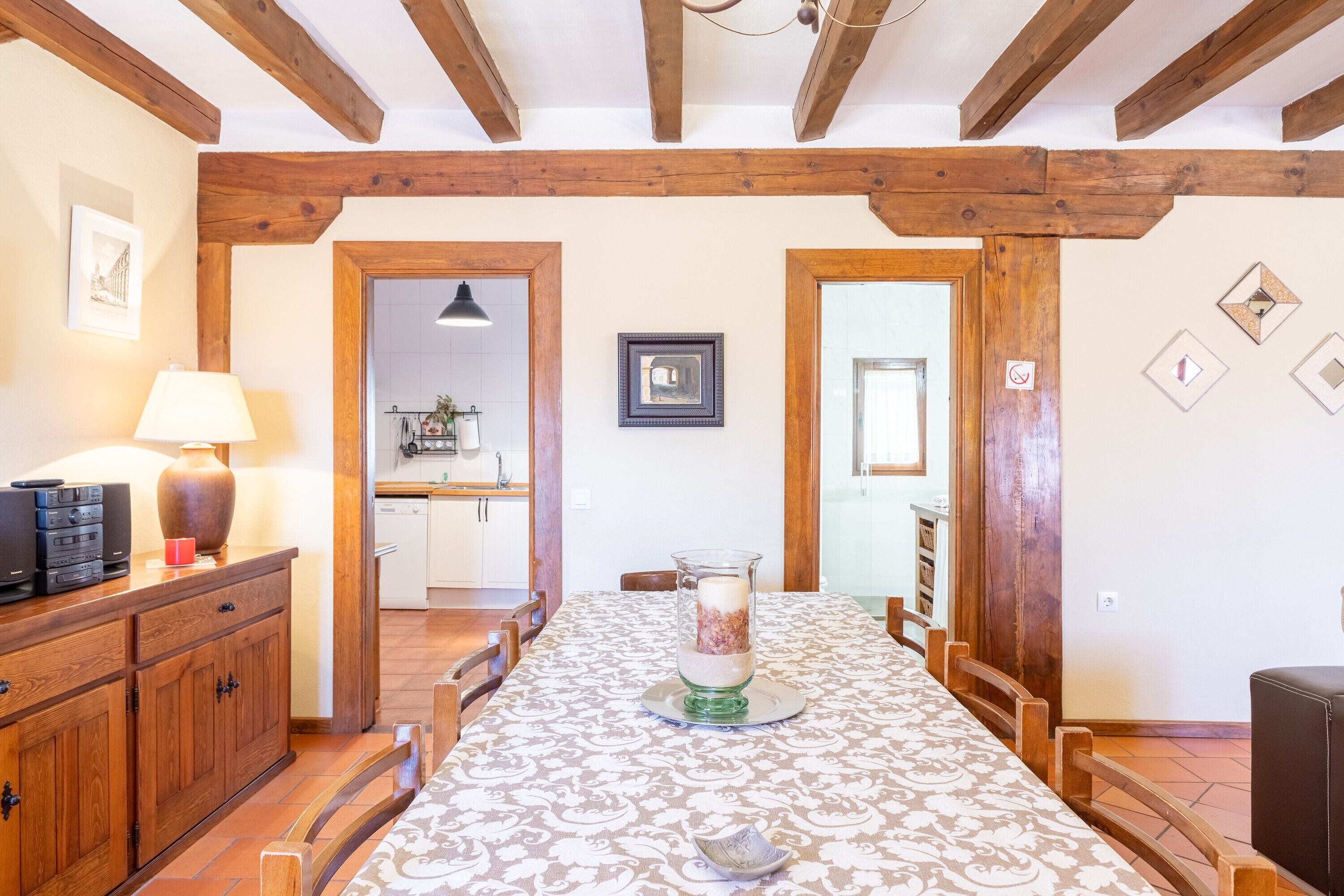 Comedor con mesa dispuesta para cuatro, sillas de madera, un florero con flores y techo de madera con vigas a la vista.