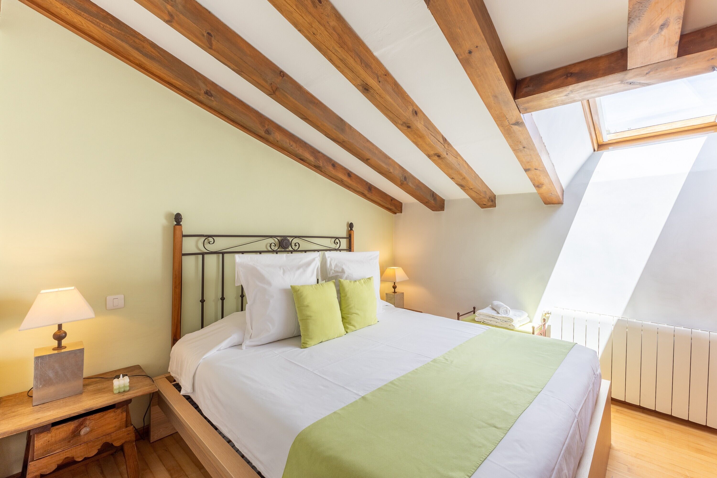 A bedroom with a bed, wooden beams, a skylight, a nightstand, and a lamp.