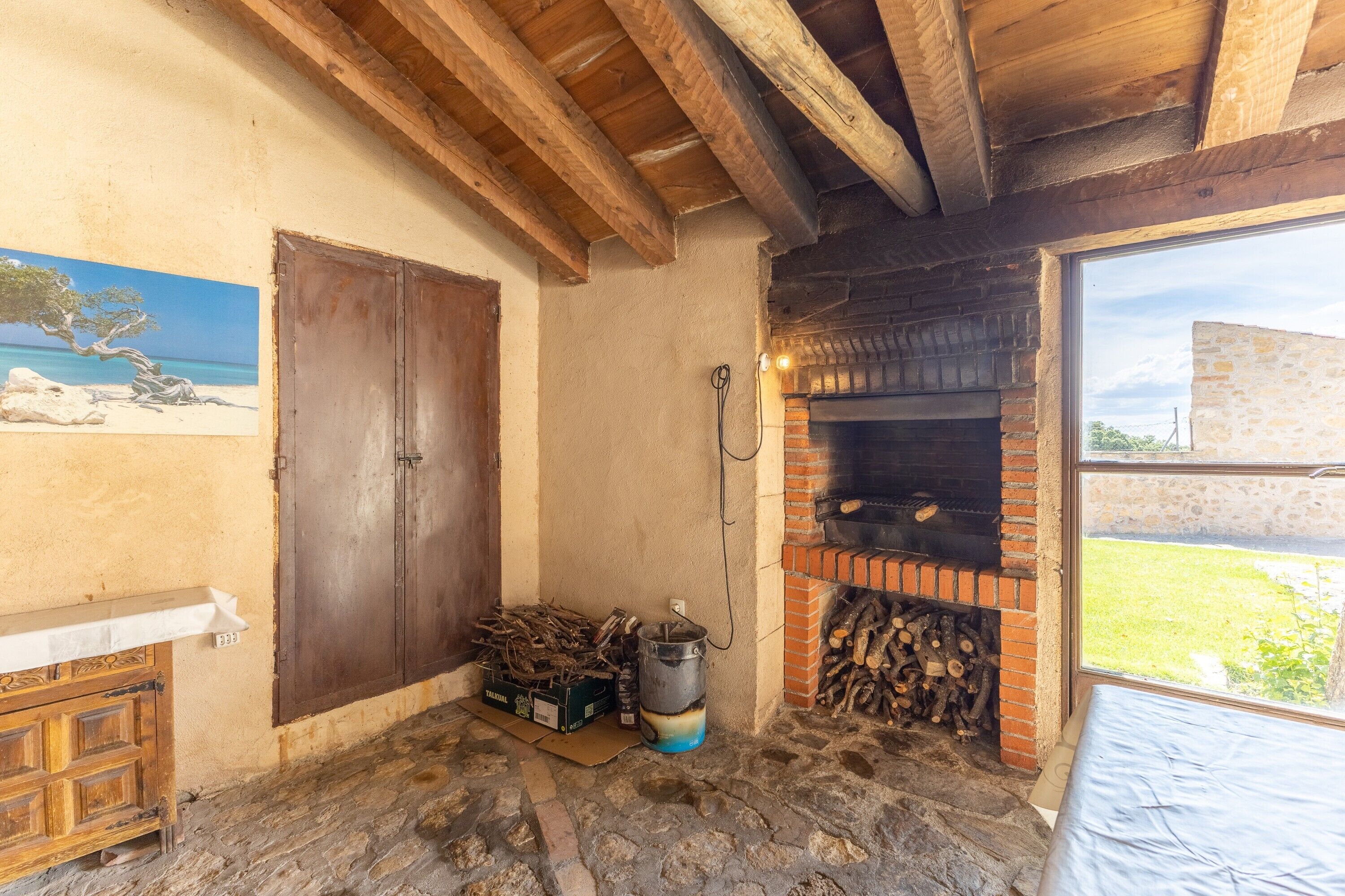 Una habitación rústica con techo de madera, chimenea de ladrillo, puerta de madera y ventana con vistas al exterior.