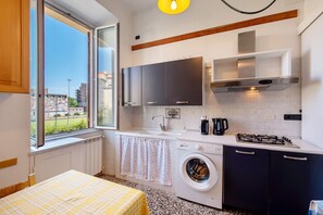 Une cuisine compacte équipée d’un lave-linge, d’une cuisinière à gaz et d’une fenêtre donnant sur des immeubles.