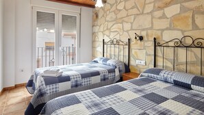 A bedroom with two beds, a stone wall, a wooden ceiling, and a balcony with shutters.