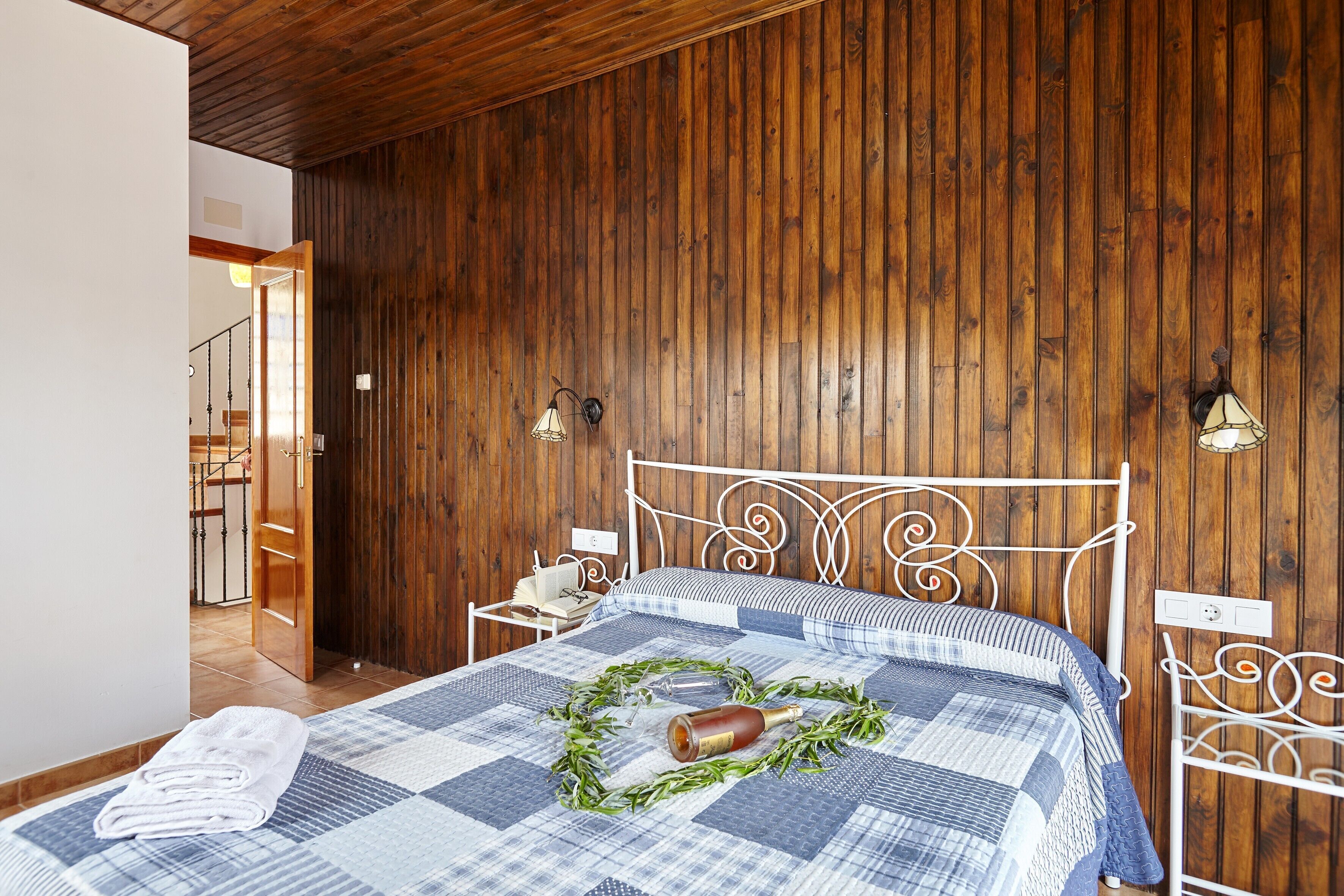 Un dormitorio con pared de paneles de madera, estructura de cama metálica, colcha acolchada y mesita de noche con lámpara.
