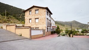A two-story stone building with a red-tiled roof, a paved driveway, and a mountainous backdrop.
