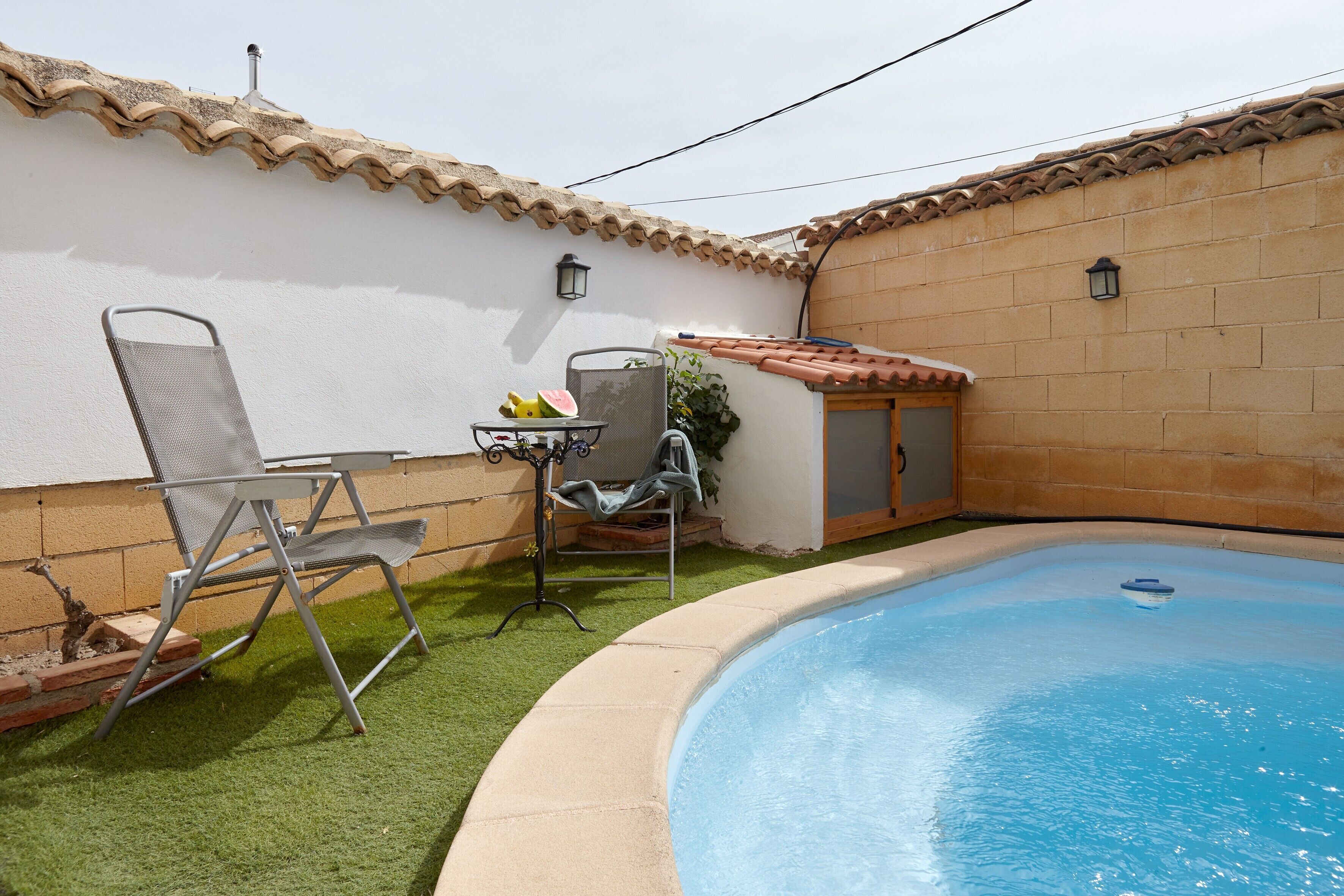 A swimming pool with a tiled roof, a chair, a small table with fruits, and a lamp post.