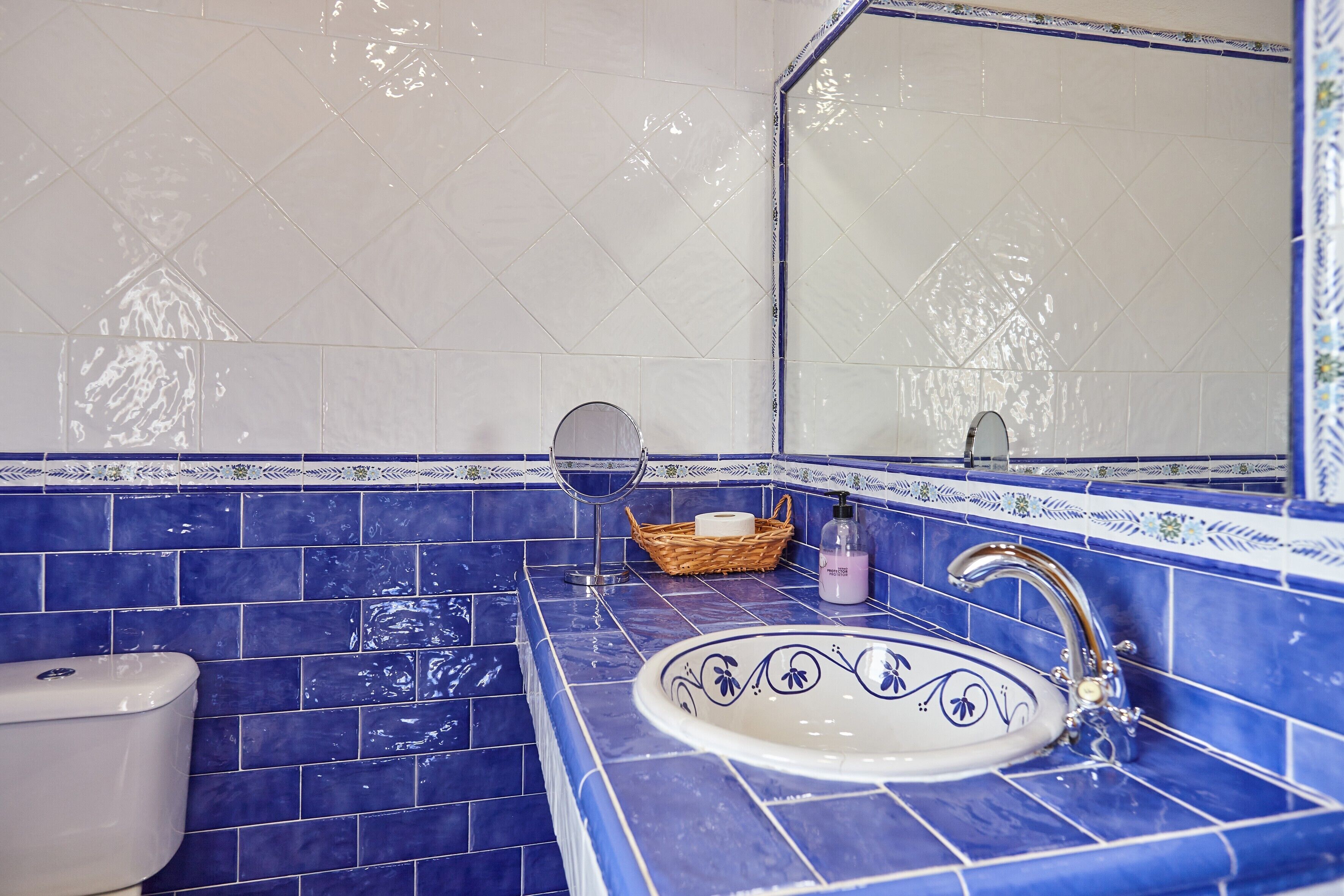 Een badkamer met blauwe tegels, een witte wastafel, een spiegel en een mand met toiletartikelen.