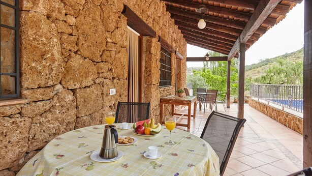 Un bâtiment rustique en pierre, doté d’un patio couvert, d’une table préparée pour le petit-déjeuner et offrant une vue sur le paysage environnant.