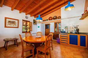 A kitchen with wooden cabinets, a dining table with chairs, and hanging utensils. - Holiday Home 'Tramuntana' with Sea View, Private Pool and Wi-Fi (Valldemossa)