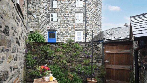 Ein Steinhaus mit einer blauen Tür, einem Holztisch mit Stühlen und einem Gartenbereich mit Pflanzen und Blumen.