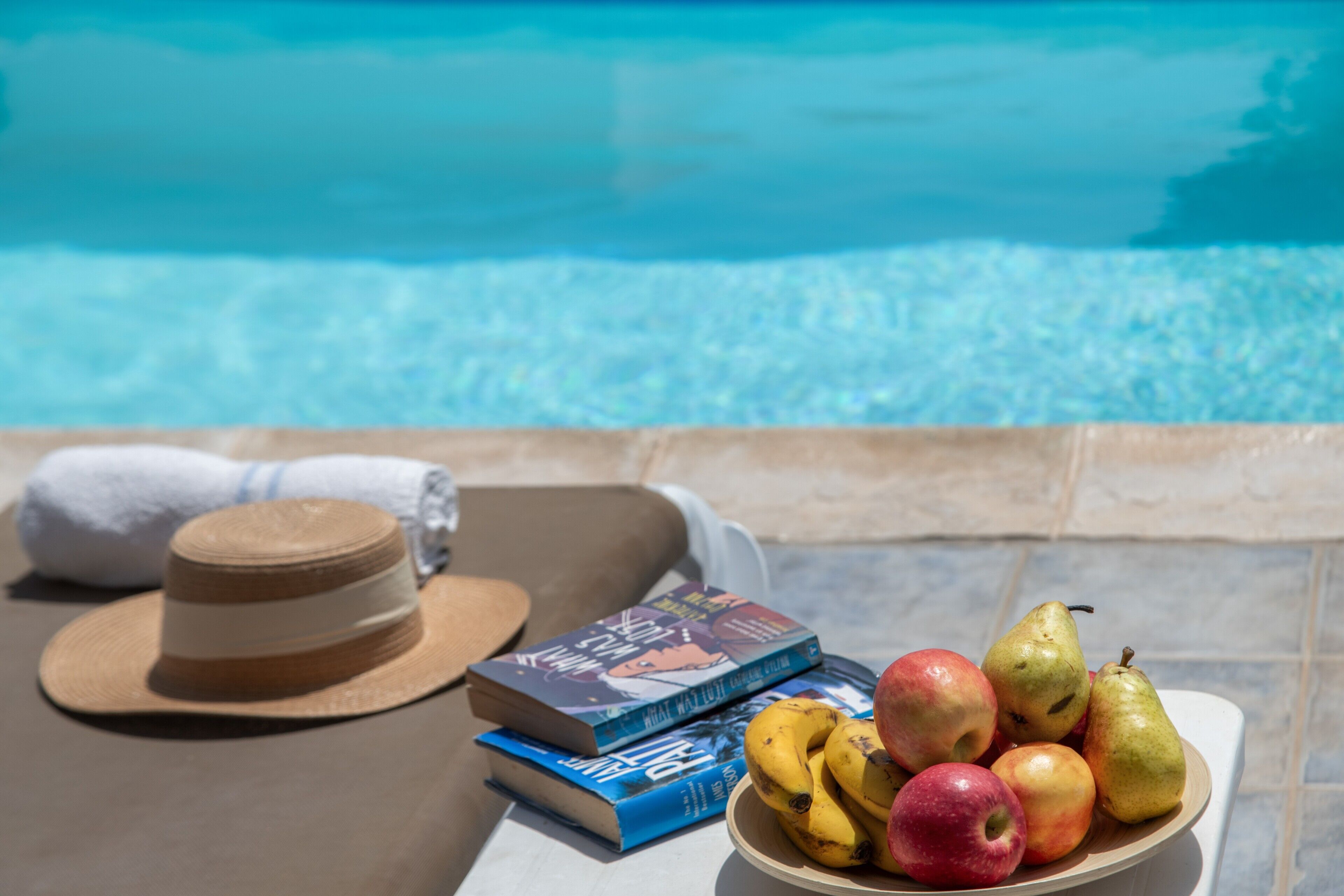 Une scène de bord de piscine avec un chapeau, des livres et un saladier rempli de fruits.