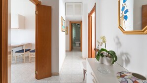Un pasillo con una puerta de madera que da a un baño, un lavamanos blanco con una planta y un espejo con un diseño floral azul.