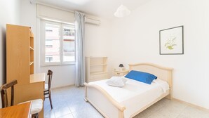 Un dormitorio con cama, escritorio, silla, armario, ventana con cortinas y lámpara de pared.