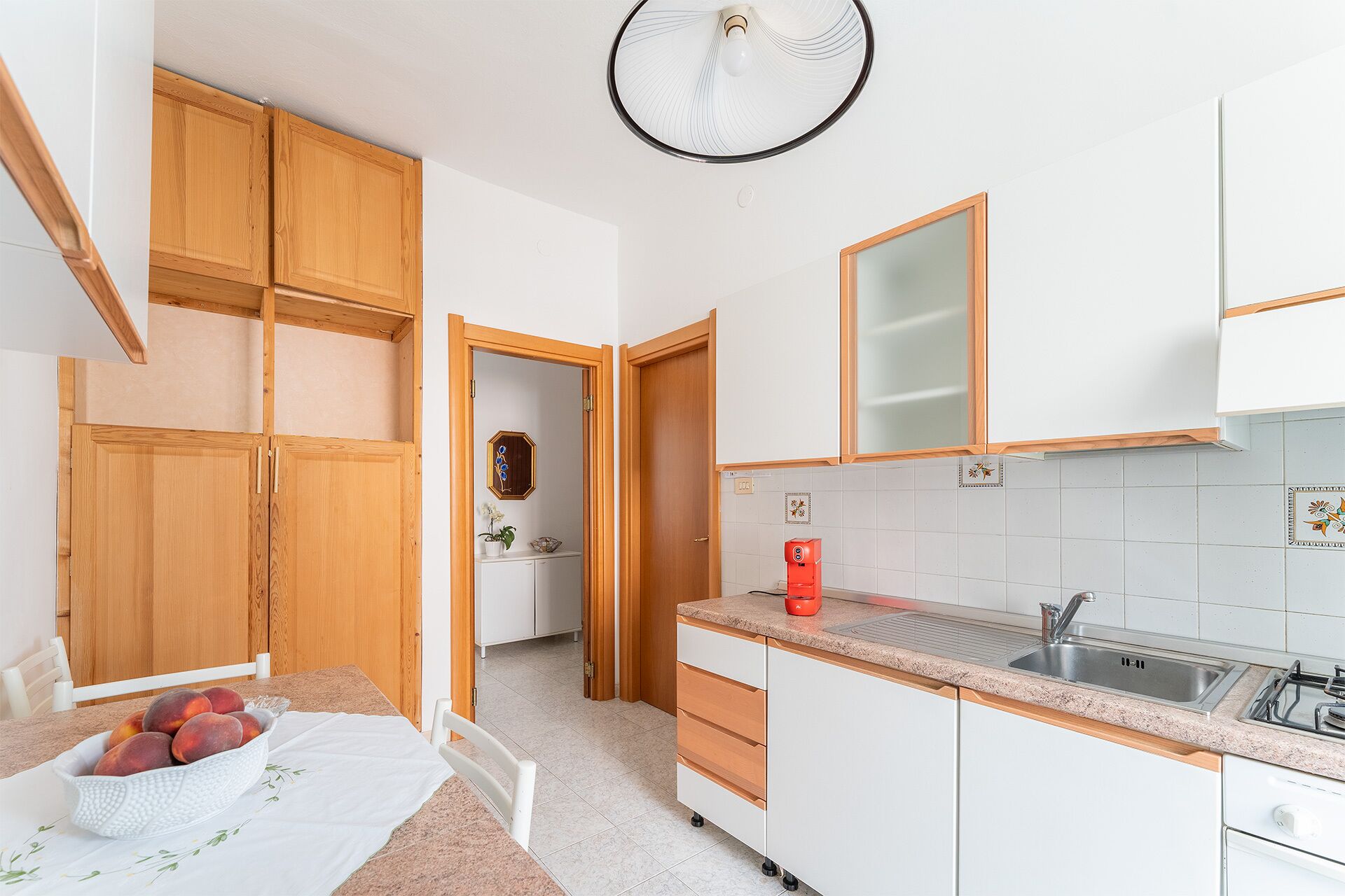 Cuina compacta amb armaris de fusta, taulell blanc, un rentaplat i ventilador al sostre.