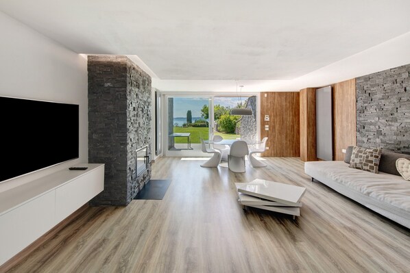 A modern living room with a flat-screen TV, a stone accent wall, a wooden floor, a dining area with white chairs, and a sofa with a patterned cushion.
