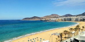 A coastal cityscape with a beach, clear blue waters, and a mountainous backdrop.