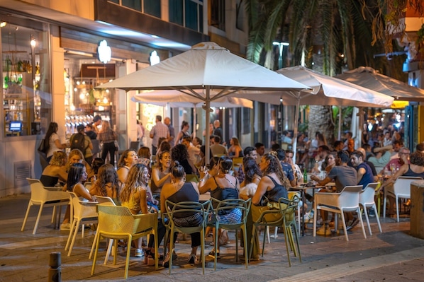 An outdoor dining area with tables and chairs, umbrellas, and a crowd of people.