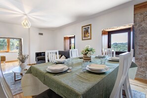 A dining area with a table set for four, white chairs, a vase with flowers, and a view of the outdoors through large windows. - Holiday Home 'Casa Do Pião - Turismo Rural' with Mountain View, Wi-Fi and Air Conditioning (Ansião)