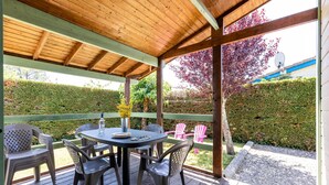 A covered outdoor patio with a wooden table and chairs, surrounded by a wooden deck and a hedge.