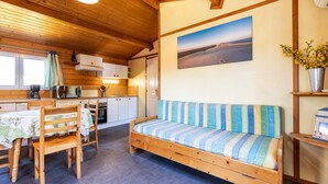 A cozy cabin interior with a wooden dining table, chairs, a sofa, a kitchen area, and a large window.
