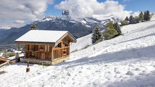 Una cabaña de madera con techo cubierto de nieve, rodeada de montañas y árboles nevados.
