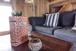 A rustic interior with a patterned container labeled 'ODES' on a wooden table, a grey sofa with striped pillows, and a decorative white vase.