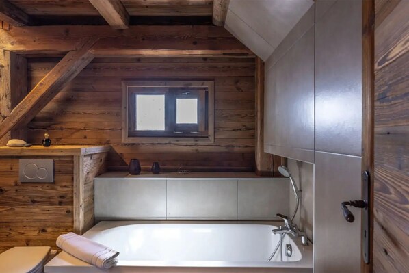 Une salle de bain rustique, située dans une cabane en bois, avec un plafond en bois, une fenêtre, une baignoire et des toilettes.