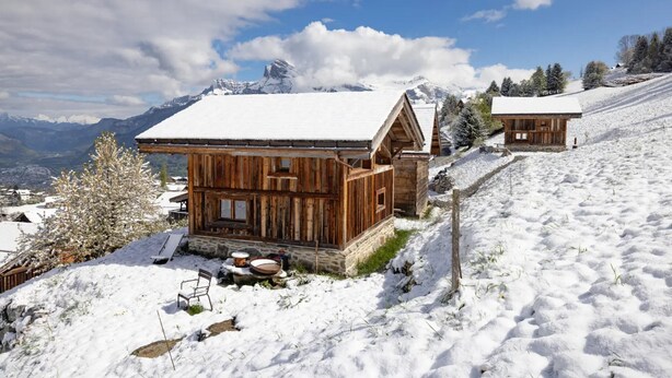 A wooden cabin with a snow-covered roof, a stone foundation, and a view of snow-covered mountains in the background. - Chalet 'Mazot Des Aravis' with Mountain View, Private Terrace and Private Garden (Combloux)