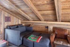 Une pièce chaleureuse avec un plafond en bois, un canapé gris, un fauteuil en cuir marron et un plaid à motifs.
