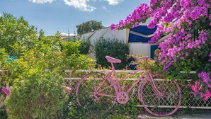 Una bicicleta rosa apoyada en una valla con flores moradas y follaje al fondo.