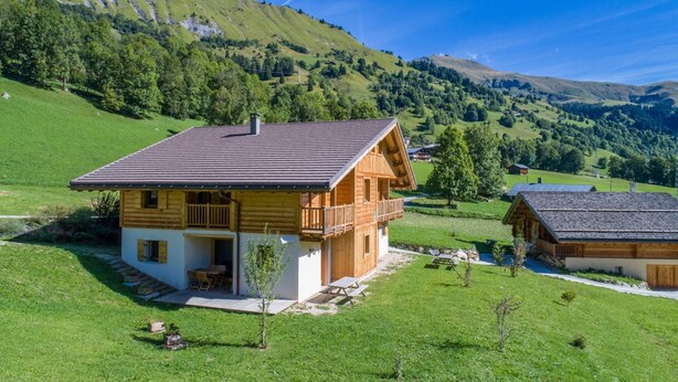 Eine Holzhütte mit Balkon, eingebettet in ein grünes Tal mit Hügeln im Hintergrund.