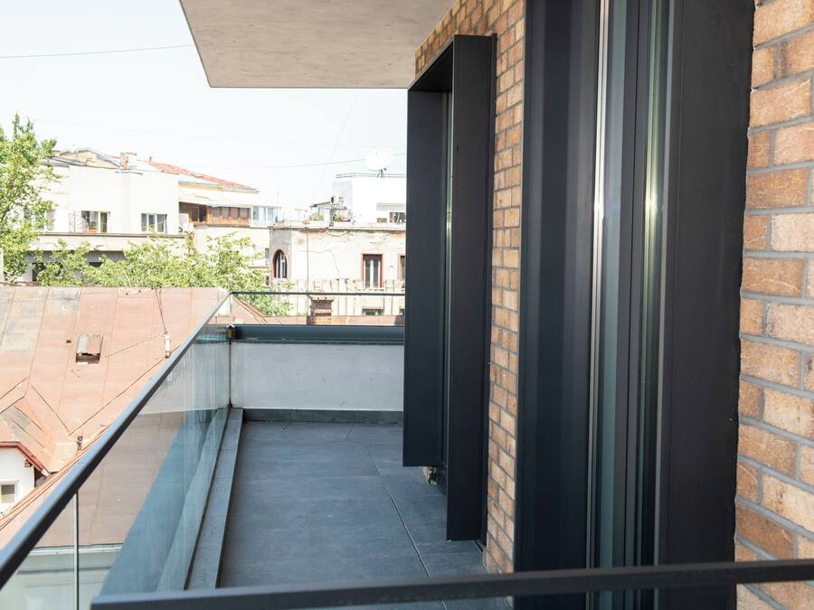 Un balcón con barandilla de vidrio, pared de ladrillo y vista a edificios vecinos.