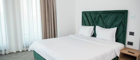 A hotel room with a large bed, green headboard, white bedding, a wooden nightstand, and a window with sheer curtains.
