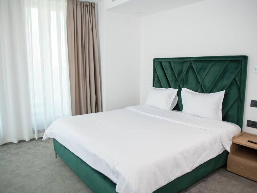 Habitación de hotel con una cama grande, cabecera verde, ropa de cama blanca, una mesita de noche de madera y una ventana con cortinas translúcidas.