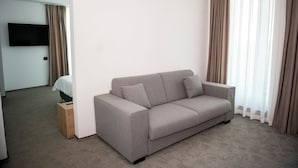 Una habitación de hotel moderna con un sofá gris, un televisor de pantalla plana montado en la pared y una cama con cabecera de madera.