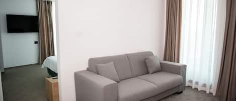 A modern hotel room with a grey sofa, a flat-screen TV mounted on the wall, and a bed with a wooden headboard.