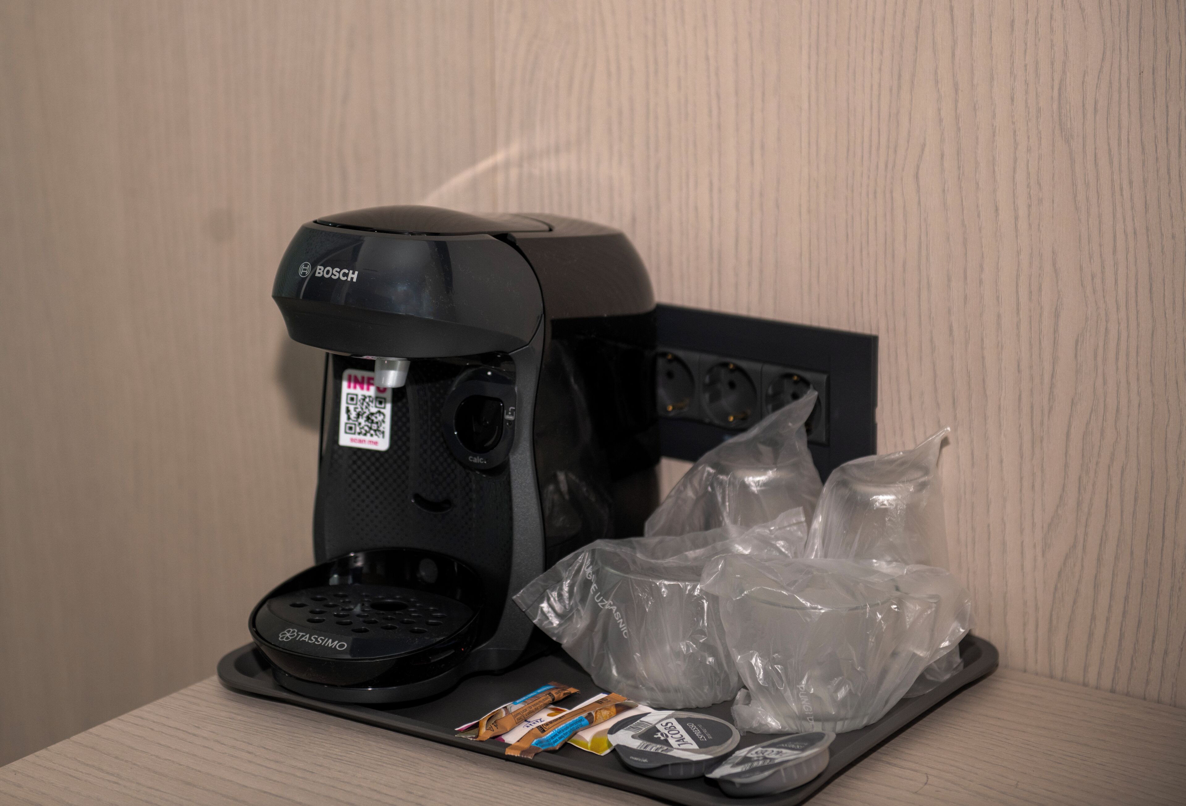 Kaffemaskine med en QR-kode, bakke med kaffekapsler og plastikposer på et bord.