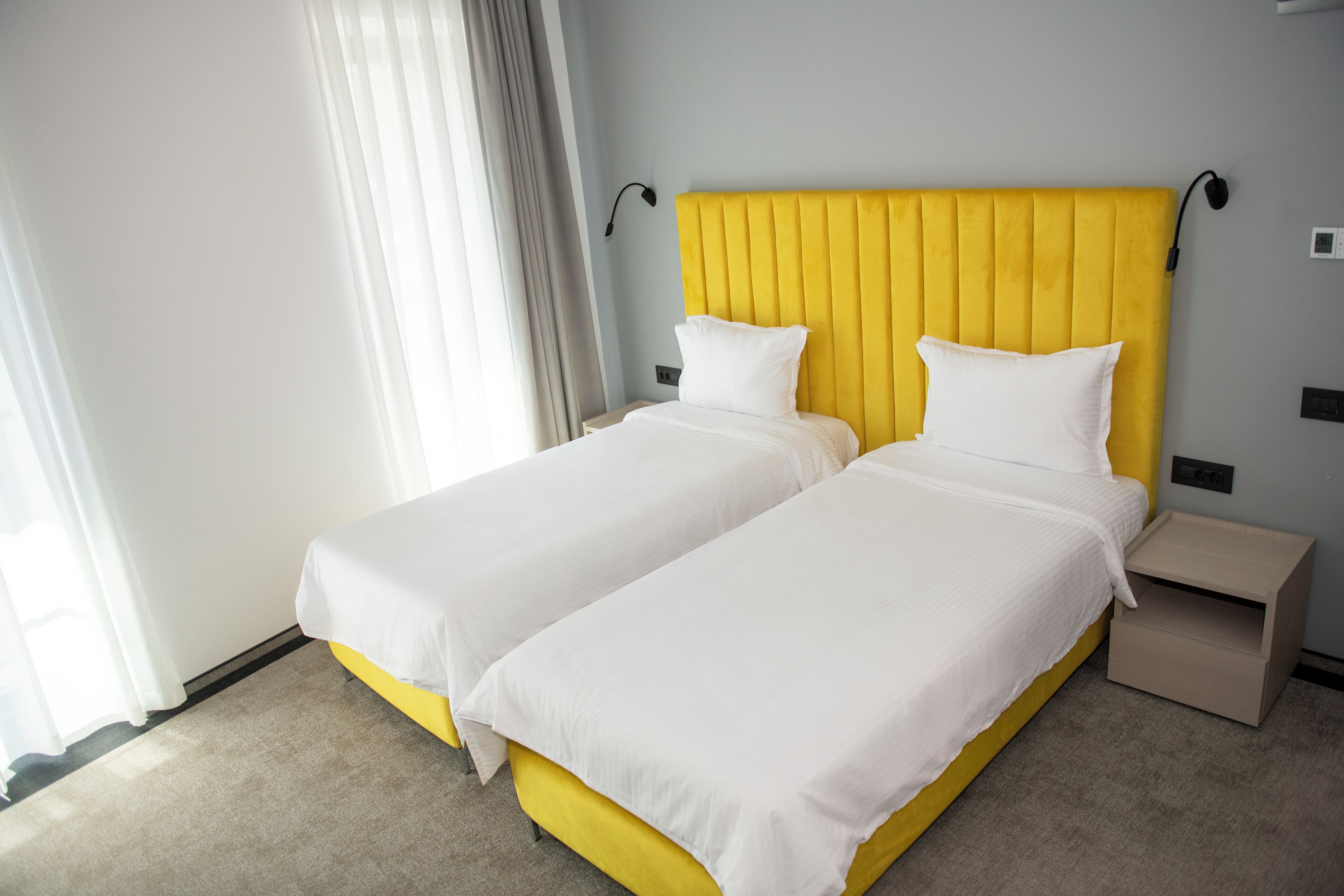 Hotelkamer met twee eenpersoonsbedden, een geel hoofdeinde, wit beddengoed, een nachtkastje en een raam met gordijnen.