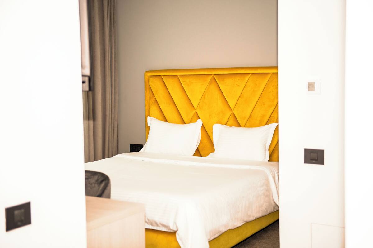 A hotel room with a yellow headboard, white bedding, and a neutral color scheme.