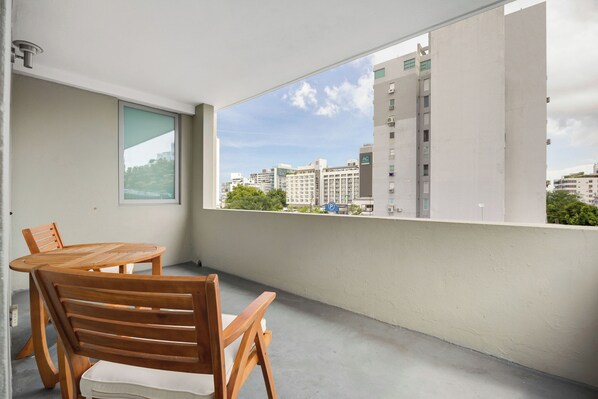 Un balcón con una mesa redonda de madera y sillas, un ventanal que ofrece vistas de edificios y vegetación circundante.